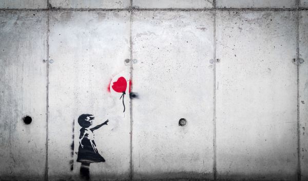 image on concrete wall of small girl with arm outstretched, reaching toward a red heart-shaped ballon that has slipped from her grasp